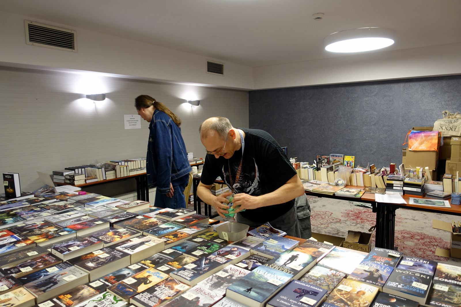 Big flat surface is covered with books. Behind it there is a man tearing the foil bag. In the background there is another man standing and there are two tables with books on them.